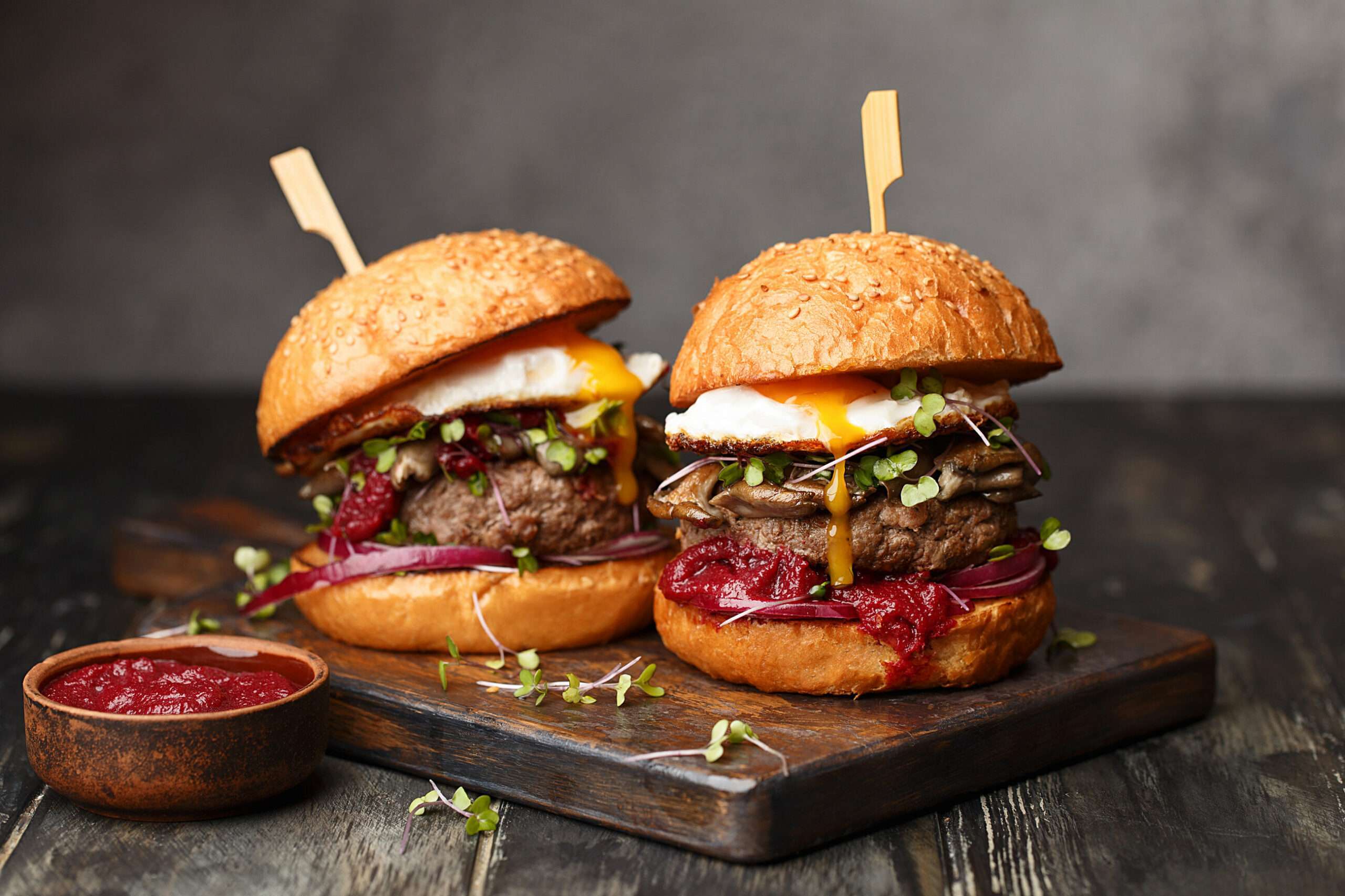 Two homemade beef burgers with mushrooms, micro greens, red onion, fried eggs and beet sauce on wooden cutting board. Side view, close up